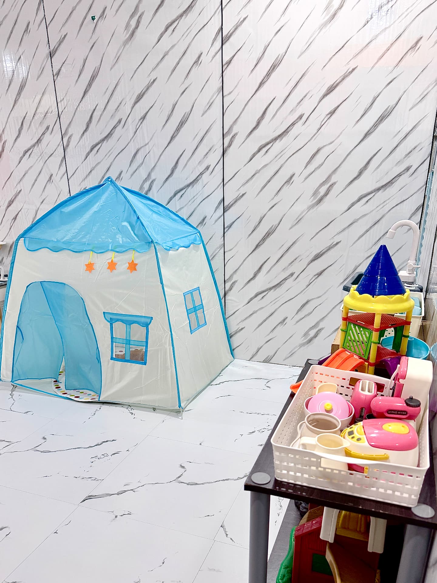 A blue and white play tent in a bright room next to a trolley of toys.