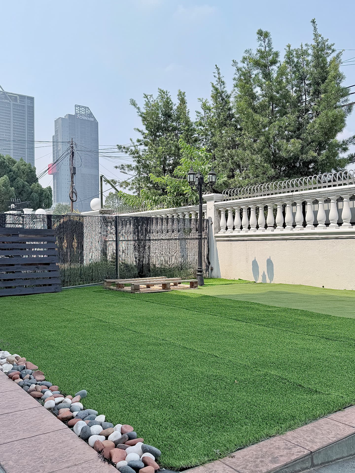 An outdoor green lawn with a wooden bench, trees, and city buildings in the background.